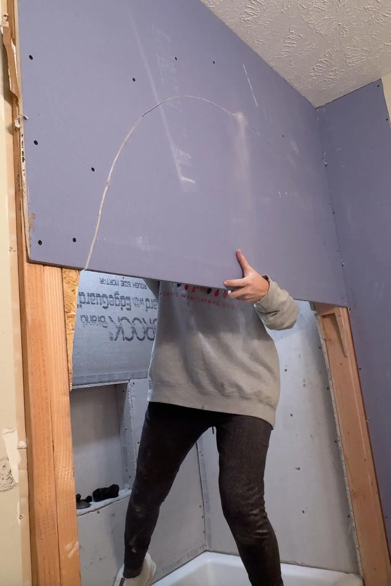 A woman holding a partially cut purple drywall sheet with an arched cutout. She is standing in a bathtub while working on attaching the sheet to an unfinished bathroom wall during a renovation project.